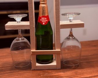 May include: A light-colored wooden caddy holds a green glass bottle with a gold foil neck and a red label reading NEW REFRESHING TASTE. Two clear glass wine glasses are suspended upside down on the side arms of the wooden frame.
