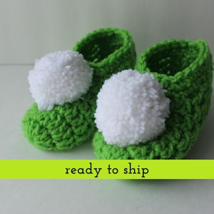 May include: A pair of handmade green crochet baby booties with white pom-poms. The booties are made of a soft yarn and are designed to keep a baby's feet warm. The text "ready to ship" is visible in the image.