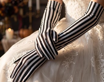 May include: A pair of long, elbow-length opera gloves featuring a bold black and white vertical striped pattern. The gloves are made of a smooth, fitted fabric that extends from the fingers up to the mid-forearm, worn over a white lace dress.