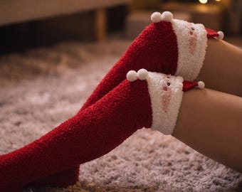 Könnte beinhalten: Ein Paar rote, flauschige Kniestrümpfe mit einem weißen Plüschbündchen, das wie ein Weihnachtsmann-Gesicht gestaltet ist. Das Bündchen zeigt eine kleine rote Mütze, gestickte Gesichtszüge und zwei weiße Bommeln am oberen Rand.