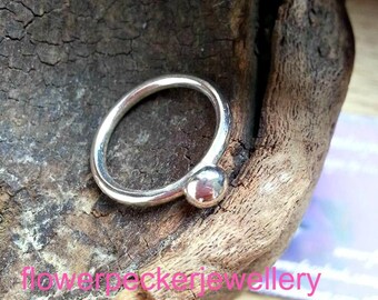 Peut inclure: Une bague en argent avec une petite boule à l'extrémité. La bague est posée sur un morceau de bois.