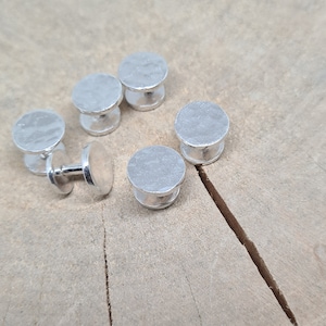 May include: Five silver cufflinks with a textured surface. The cufflinks are arranged in a semi-circle on a wooden surface.