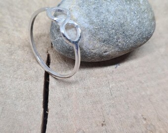 Peut inclure: Bague en argent avec un symbole infini. La bague est posée sur une surface en bois à côté d'une pierre grise. Le symbole infini est au centre de la bague.