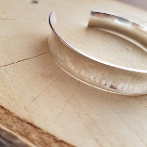 May include: A silver bracelet with a hammered texture. The bracelet is wide and has a rounded shape.