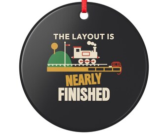May include: A round black ornament with a red ribbon hanger featuring a graphic of a white steam train on tracks, a green hill, and the text THE LAYOUT IS NEARLY FINISHED in white and gold lettering centred on the surface.