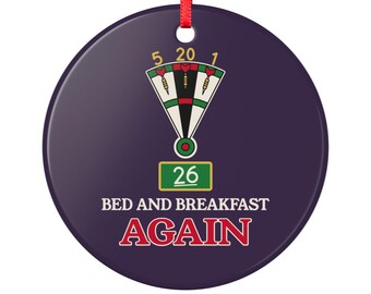 May include: A round dark purple ornament with a red ribbon hanger. The design features a dartboard segment with numbers 5, 20, and 1, a green box with the number 26, and the white and red text "BED AND BREAKFAST AGAIN" printed below.