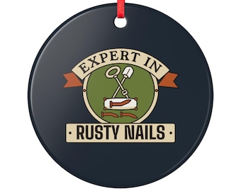 May include: A round dark blue ornament with a red ribbon hanger. The centre features a green circle with a shovel and metal detector icon, and three rusty nails. The text reads EXPERT IN RUSTY NAILS in a beige banner and rectangular box.