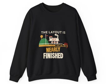 May include: A black long-sleeve sweatshirt featuring a graphic of a small train on tracks with a green hill and the text THE LAYOUT IS NEARLY FINISHED in white and yellow lettering centred on the front of the garment.