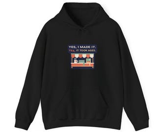 May include: A black hooded sweatshirt featuring a graphic print of a market stall with a coffee mug and a computer. The text above the stall reads, YES, I MADE IT. YES, IT TOOK AGES. The hoodie has long sleeves and a front kangaroo pocket.