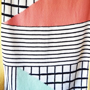 Peut inclure: Une couverture en tricot avec un motif géométrique en noir, blanc, corail et vert menthe. La couverture a une bordure jaune.