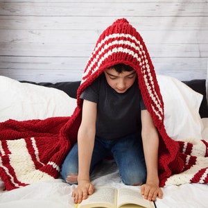 May include: A child wearing a red and white striped hooded blanket sits on a white bed and reads a book.