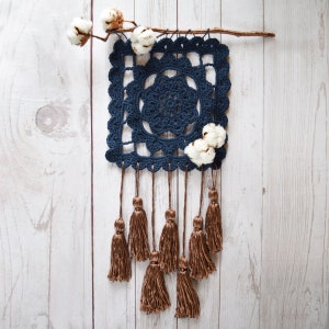 Può includere: Un arazzo da parete quadrato in crochet blu navy con una frangia a nappe. L'arazzo presenta un motivo geometrico ed è decorato con boccioli di cotone.