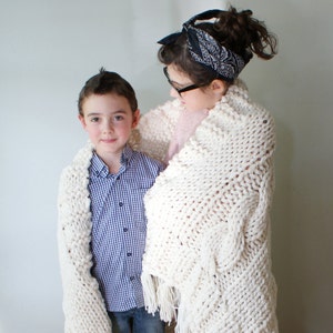 May include: A large, cream-colored, chunky knit blanket with fringe, draped over two people. The blanket has a textured pattern and is made of thick yarn. The blanket is a cozy home decor item.