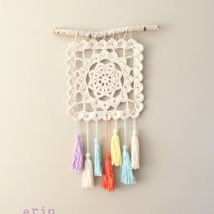 May include: A white crocheted square wall hanging with a floral design and colorful tassels in shades of purple, blue, yellow, and orange. The hanging is attached to a natural branch.