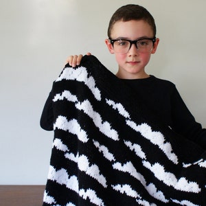 May include: A child wearing black-framed glasses holds a black and white striped blanket with a cloud pattern.