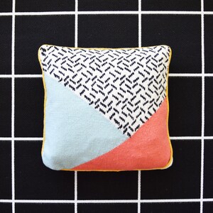May include: A geometric patterned pillow with a black and white chevron design, a light blue section, and a coral pink section. The pillow has a yellow border.
