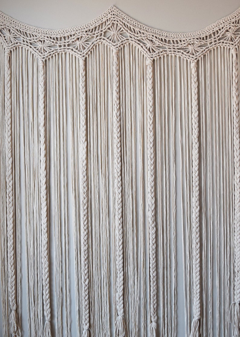 May include: A white macrame wall hanging with a scalloped top and long strands of knotted cord.