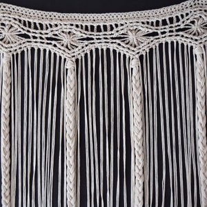 May include: A white macrame wall hanging with a geometric pattern and fringe. The hanging is made of cotton cord and has a natural, bohemian style.