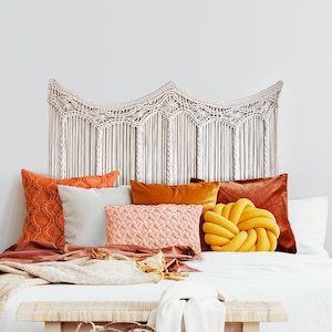 May include: A white macrame headboard with a wavy design hangs above a bed with a white duvet, brown throw blanket, and several pillows in shades of orange, yellow, and pink.