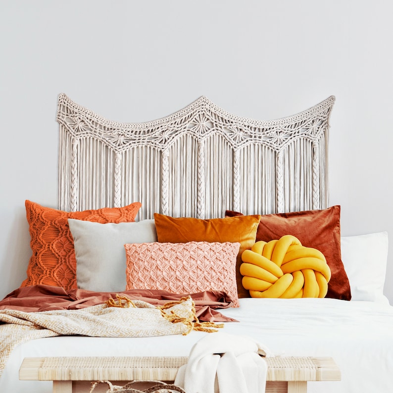 May include: A white macrame headboard with a wavy design hangs above a bed with a white duvet, brown throw blanket, and several pillows in shades of orange, yellow, and pink.