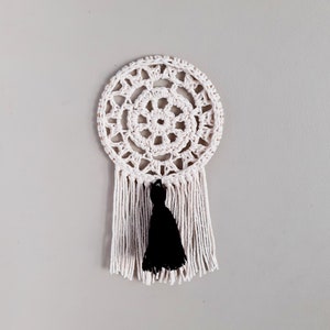 May include: White crocheted dream catcher with a black tassel hanging from the bottom. The dream catcher has a circular design with a smaller circle in the center.