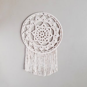 May include: A white crocheted dreamcatcher with a circular design and fringe. The dreamcatcher has a delicate pattern of flowers and stars.