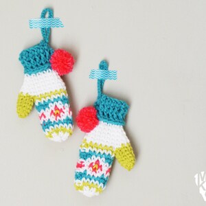 May include: Two crocheted mitten ornaments with a white, green, blue, and pink striped pattern. Each mitten has a pink pom-pom on top.