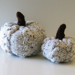 May include: Two knitted pumpkins, one larger than the other, are made with a white yarn with flecks of brown and gray. The pumpkins have brown stems.