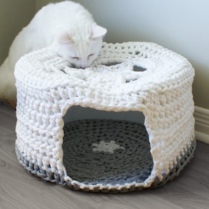 May include: A white crocheted cat bed with a gray crocheted bottom. The bed has a round opening and a white cat is looking inside.