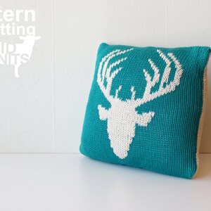 May include: A teal blue knitted pillow with a white deer head design. The pillow is on a white surface.