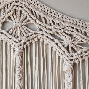 May include: A beige macrame wall hanging with a geometric pattern and long fringe. The hanging is made with thick cotton cord and has a delicate, intricate design.
