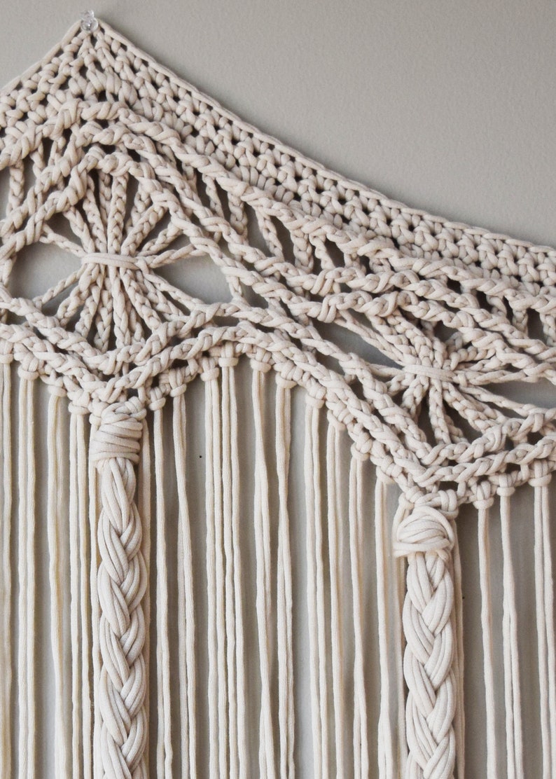 May include: A beige macrame wall hanging with a geometric pattern and long fringe. The hanging is made with thick cotton cord and has a delicate, intricate design.