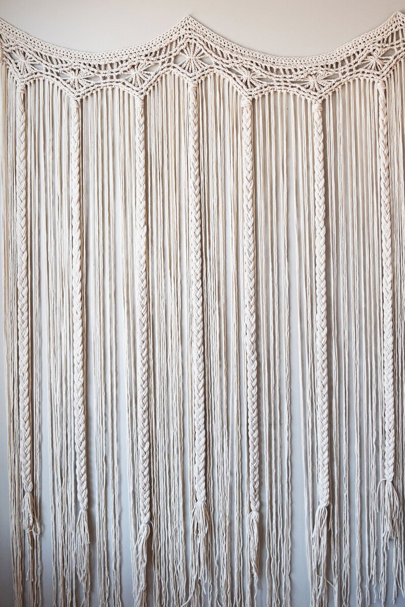 May include: A white macrame wall hanging with a scalloped top and long, braided strands.