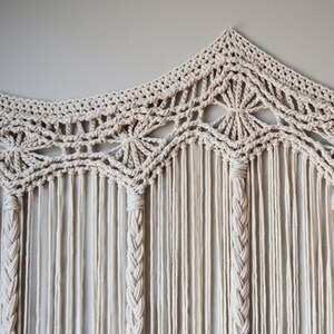 May include: A white macrame wall hanging with a geometric design. The hanging is made of cotton cord and has a fringe at the bottom.