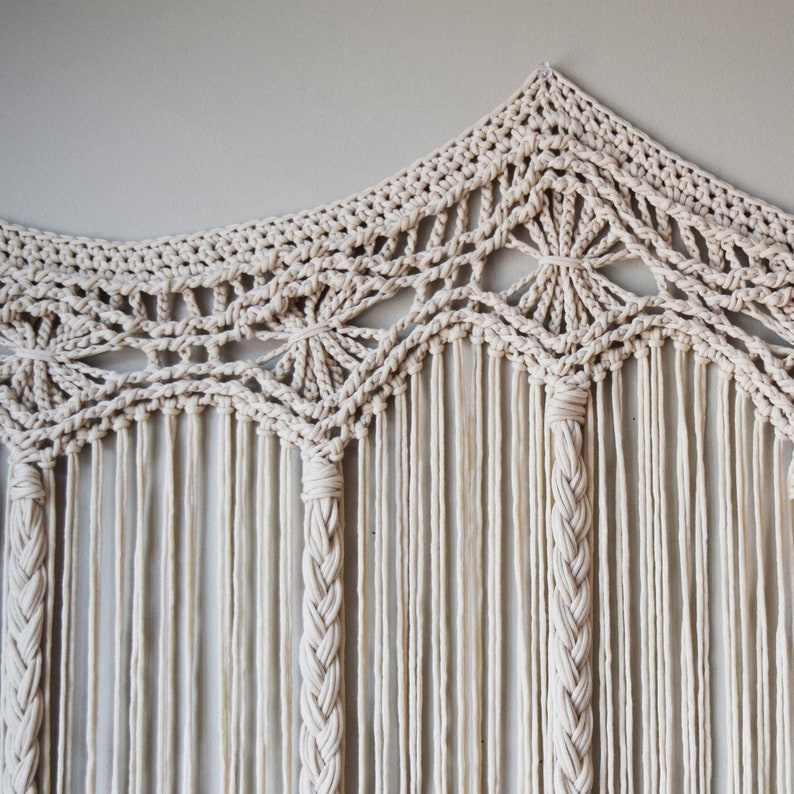 May include: A white macrame wall hanging with a geometric design. The hanging is made of cotton cord and has a fringe at the bottom.