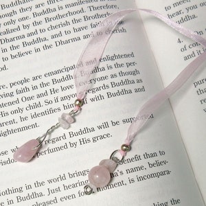 May include: A pink ribbon bookmark with two decorative charms. Each charm features a cluster of pink rose quartz beads and silver-colored wire. The bookmark is resting on the open pages of a book.