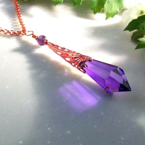 May include: A copper-colored necklace with a faceted, elongated purple crystal pendant. The pendant is suspended from a delicate chain and features intricate filigree detailing. A small purple bead accents the chain near the pendant.