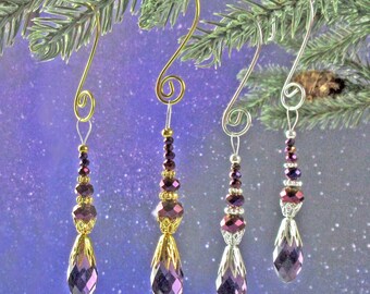 May include: Four purple and gold beaded Christmas ornaments with crystal teardrop accents hanging from a fir tree branch.