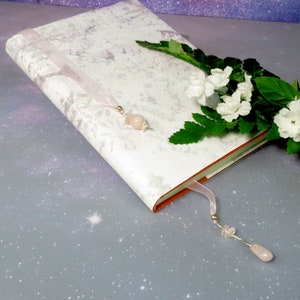 May include: A hardcover journal with a floral pattern cover, a pink ribbon bookmark, and a rose quartz charm. White flowers and green leaves are arranged on the side. The journal's pages have orange edges.
