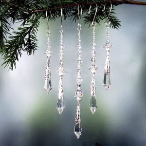 May include: Five clear crystal ornaments with silver-colored metal accents hang from a pine branch. Each ornament has a unique design, including faceted crystals and decorative metalwork. The ornaments are suspended by silver hooks.