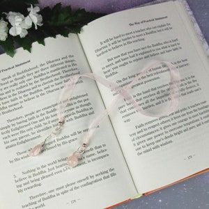 May include: A pink ribbon bookmark with two decorative charms featuring pink gemstones and silver-colored beads, resting on the open pages of a book. The book's text discusses Buddhist teachings.