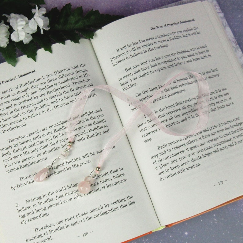 May include: A pink ribbon bookmark with two decorative charms featuring pink gemstones and silver-colored beads, resting on the open pages of a book. The book's text discusses Buddhist teachings.
