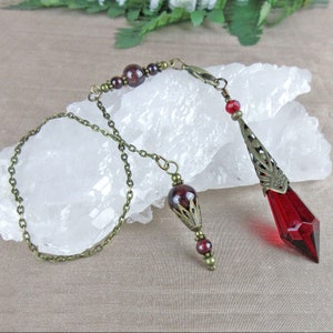 May include: A pendulum featuring a faceted deep red crystal point encased in an ornate bronze-toned metal cap. The chain includes dark red spherical beads, smaller bronze-toned accents, and a lobster clasp, connecting to a smaller matching bead charm.