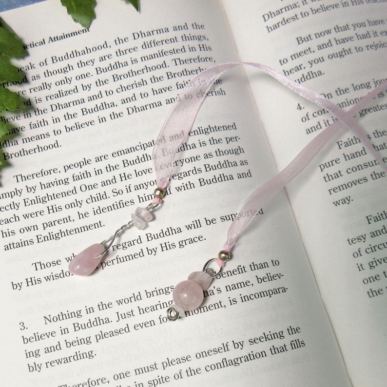 May include: A pink ribbon bookmark with two decorative charms. Each charm features a cluster of pink rose quartz beads and silver-colored wire. The bookmark is resting on the open pages of a book.