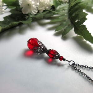 May include: A red pendant necklace with a teardrop-shaped bead and a filigree metal cap. The necklace features a dark metal chain and is set against a white background with greenery.