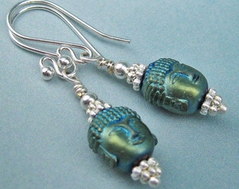 May include: A pair of silver dangle earrings with green and blue Buddha head charms. The charms are surrounded by silver beads.