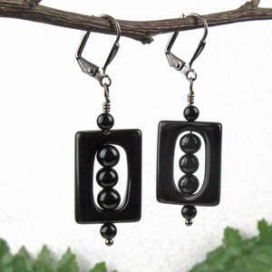 May include: A pair of black rectangular earrings with three black beads inside each rectangle. The earrings have silver-colored lever-back closures and are suspended from a thin, dark branch against a white background.