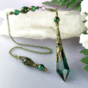 May include: A green crystal pendulum with a bronze chain and beads. The pendulum is a pointed teardrop shape with a decorative bronze top. The chain has a bronze clasp and is adorned with green tiger's eye beads.