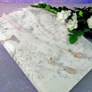 May include: Two rose quartz gemstone bookmarks with silver-colored wire and gold-colored bead accents. The bookmarks have sheer pink ribbons attached. The bookmarks rest on a floral patterned paper.