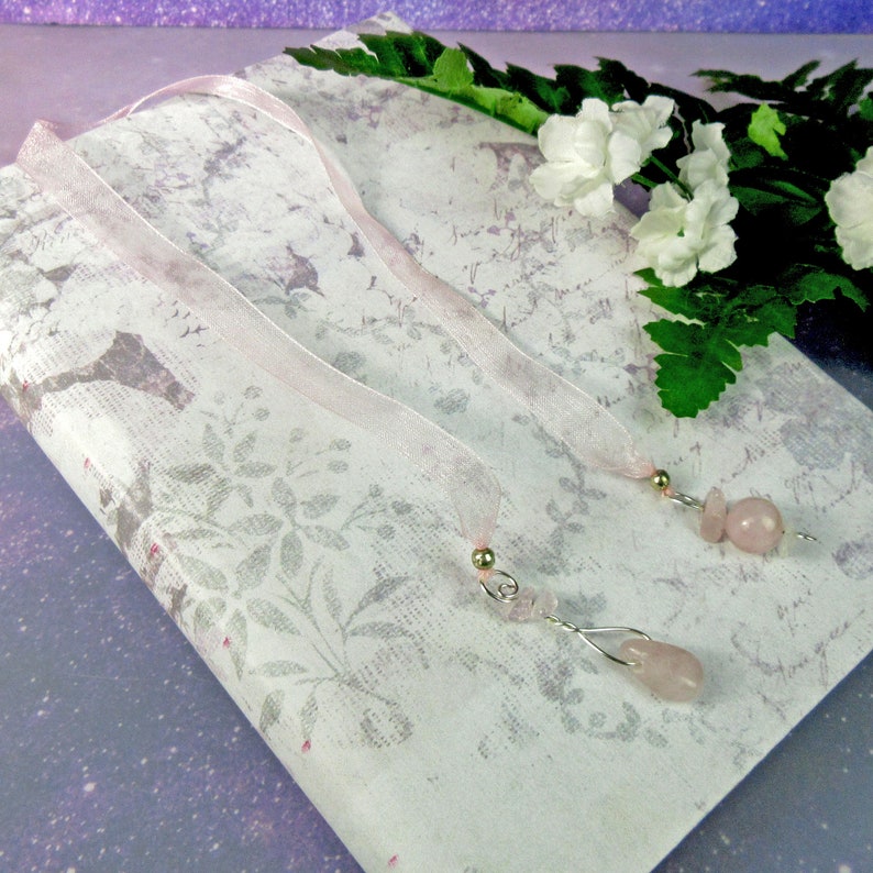 May include: Two rose quartz gemstone bookmarks with silver-colored wire and gold-colored bead accents. The bookmarks have sheer pink ribbons attached. The bookmarks rest on a floral patterned paper.